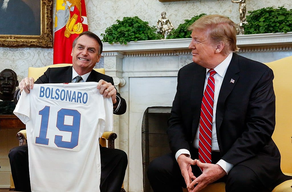 Jair Bolsonaro holds up a sports jersey that says "Bolsonaro 19" while sitting next to Donald Trump at the White House.