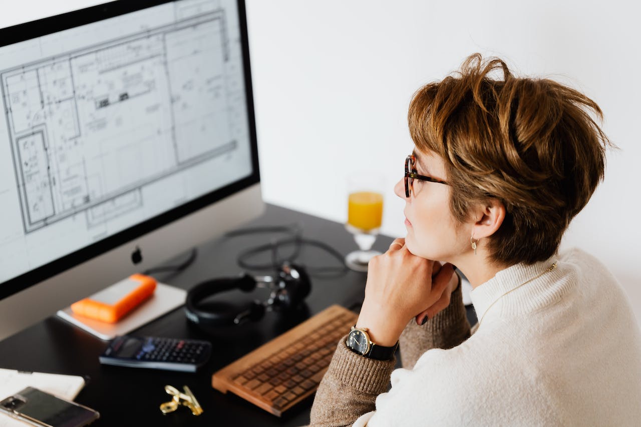 Woman reviewing blueprints on her computer monitor at her desk; image by Karolina Grabowska, via Pexels.com.