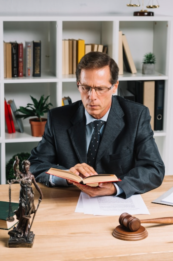 Mature male lawyer reading book with gavel and justice statue on wooden table; image by Freepik, via Freepik.com.
